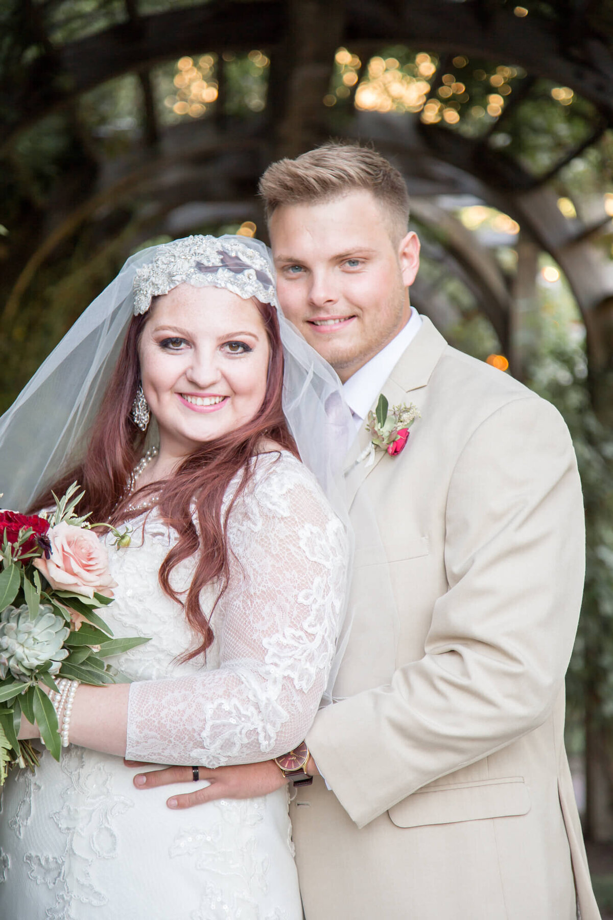 Redheaded bride standing with groom in cream suit under a garden tunnel