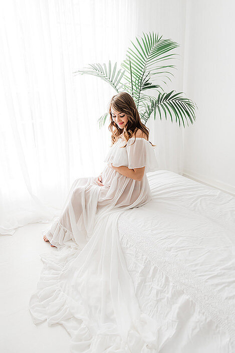 Expecting mom in an off-shoulder white gown sits by a window with a tall green palm behind her, creating a fresh and natural feel.