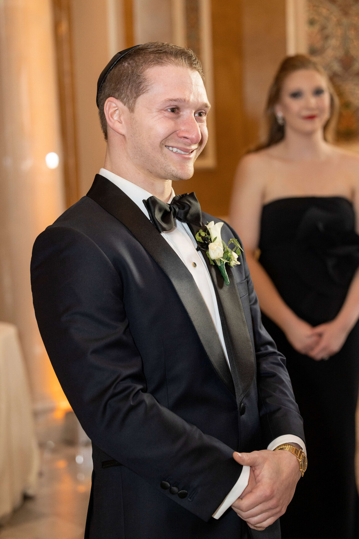 A groom in a black tuxedo with a white boutonniere smiles, standing in an elegant setting. A woman in a black dress is blurred in the background.
