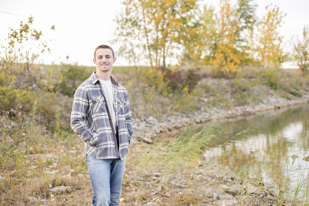 high school senior male, standing next to a pond, with trees in the background, thumbs in his flannel jacket pockets, wearing a light tan long sleeve shirt, blue jeans, looking away at the camera smiling