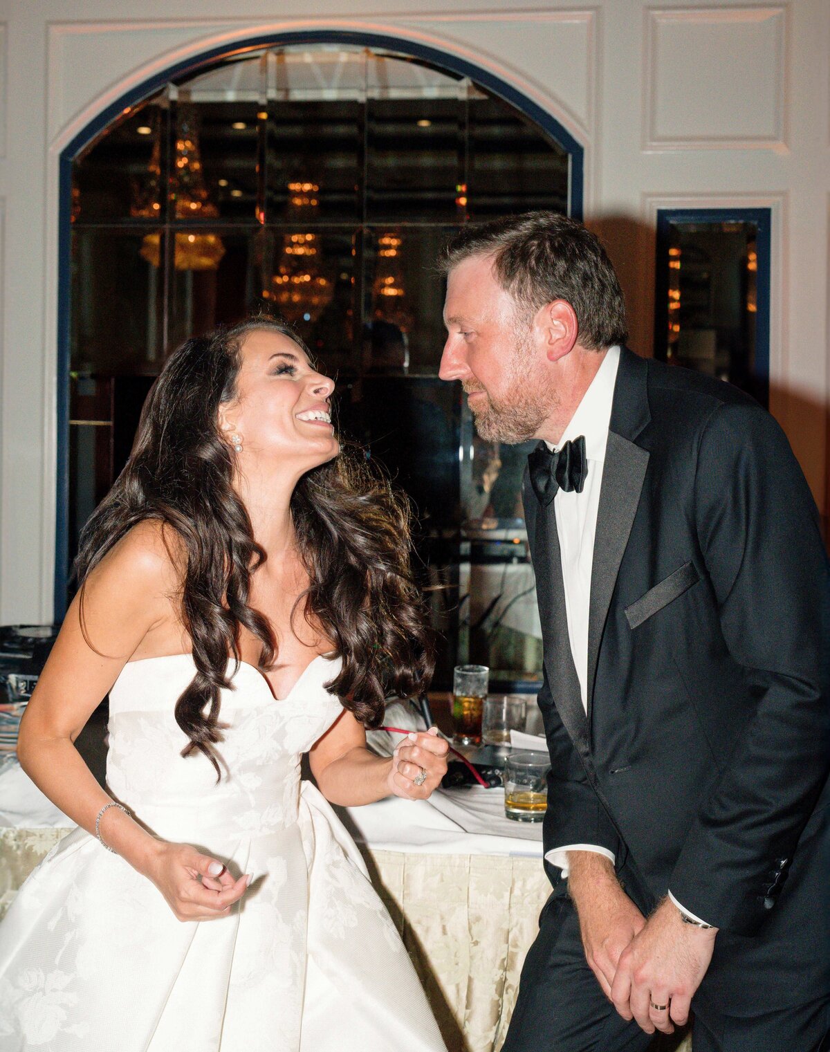 A woman in a white dress and a man in a black tuxedo share a joyful moment, smiling and laughing with each other in an elegant indoor setting.