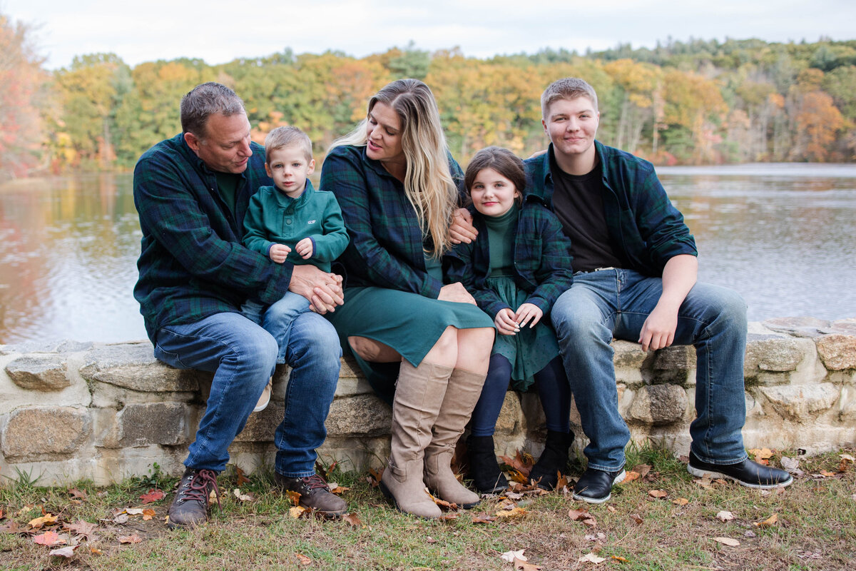 Family of five snuggled together matching in blue and green close while sitting on a rock wall by the water in the fall