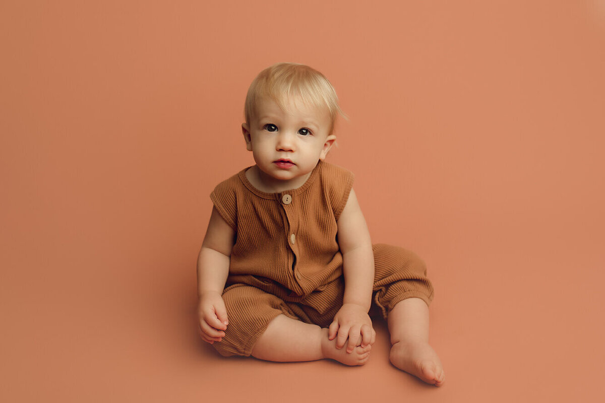 a one year old boy sitting on a tan drop wearing a tan outfit
