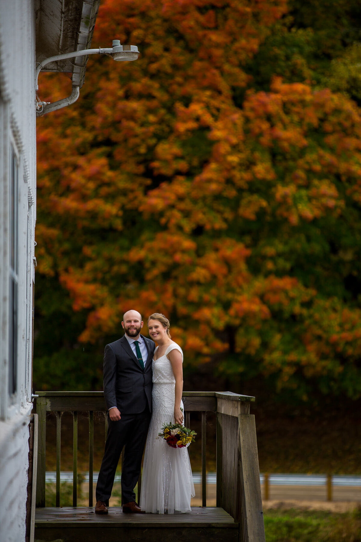 bride and groom stand on deck in front of fall foliage smiling at camera