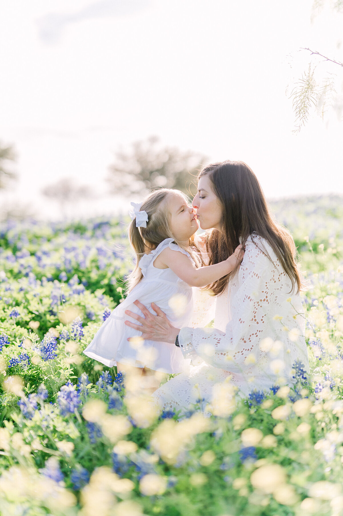 Mother delightfully kissing her toddler in a field of bluebonnets wearing all white.