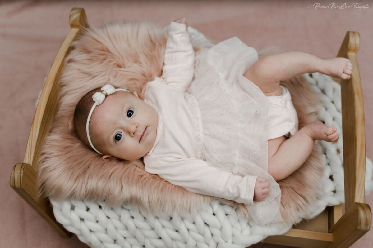 Baby girl in a soft pink dress and matching bow headband lying on a wooden newborn bed with a fluffy peach blanket.