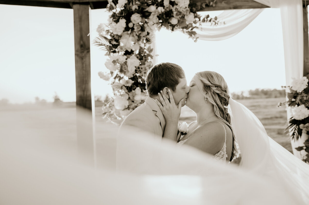 bride and groom kissing under their wedding arch as the bride holds the grooms cheeks and her veil flies in the wind