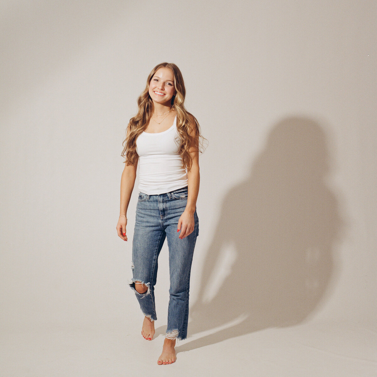 A blonde high school senior in a white top and jeans walks and laughs in a white studio