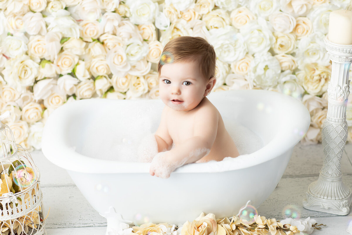 DFW milestone photographer highlights a baby sitting in a white clawfoot tub filled with bubbles, surrounded by cream roses and elegant decor, capturing a post-cake smash bath with timeless charm.