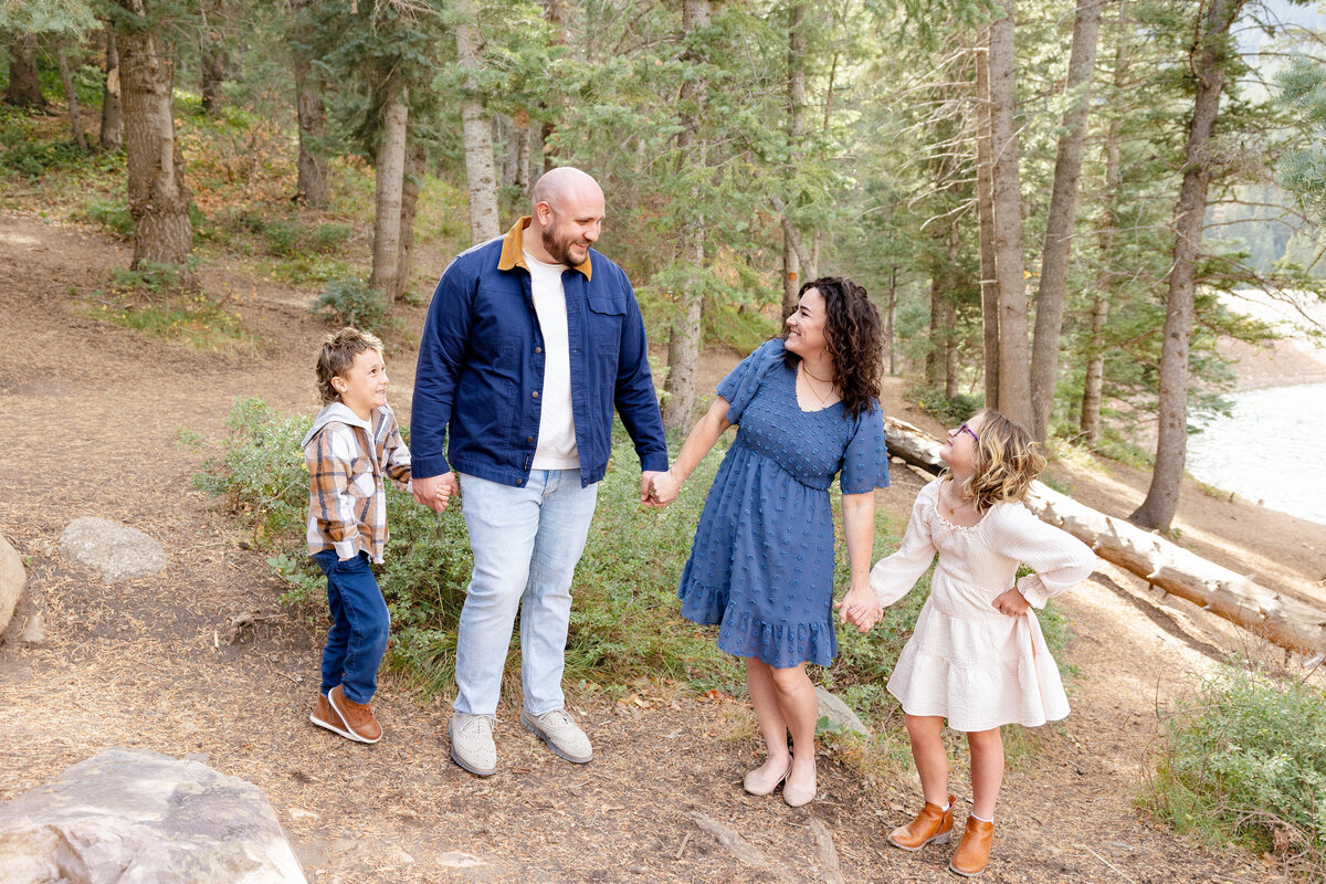  A father, mother, and their two children hold hands as they enjoy a playful walk along a forested path at Tibble Fork Reservoir, with lush greenery surrounding them.