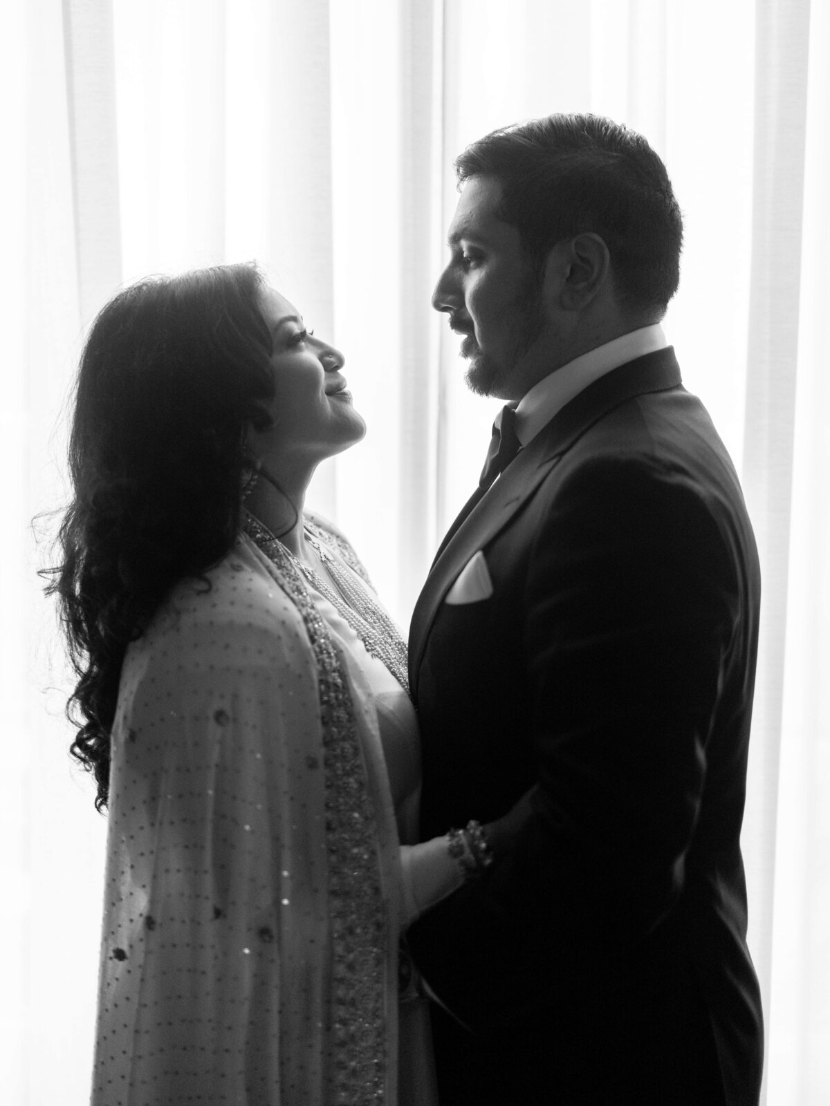 A couple stands facing each other in front of a softly lit window. The woman is wearing a traditional embellished outfit, and the man is in a suit and tie. Both are smiling affectionately. The photo is in black and white.