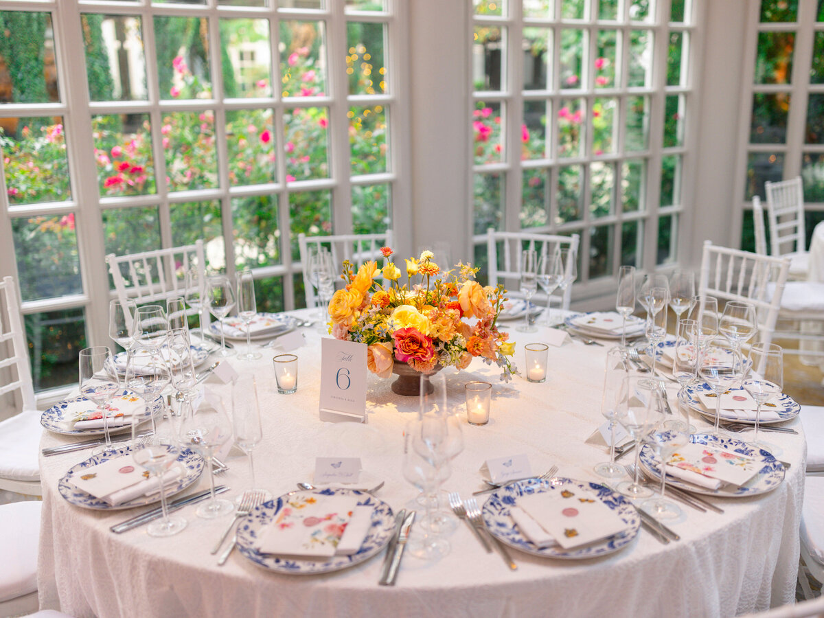 A round table set for an elegant event, featuring white chairs and a white tablecloth. The centerpiece is a vibrant floral arrangement with yellow and orange flowers. Each place setting includes a blue and white patterned plate, cutlery, and a wine glass.