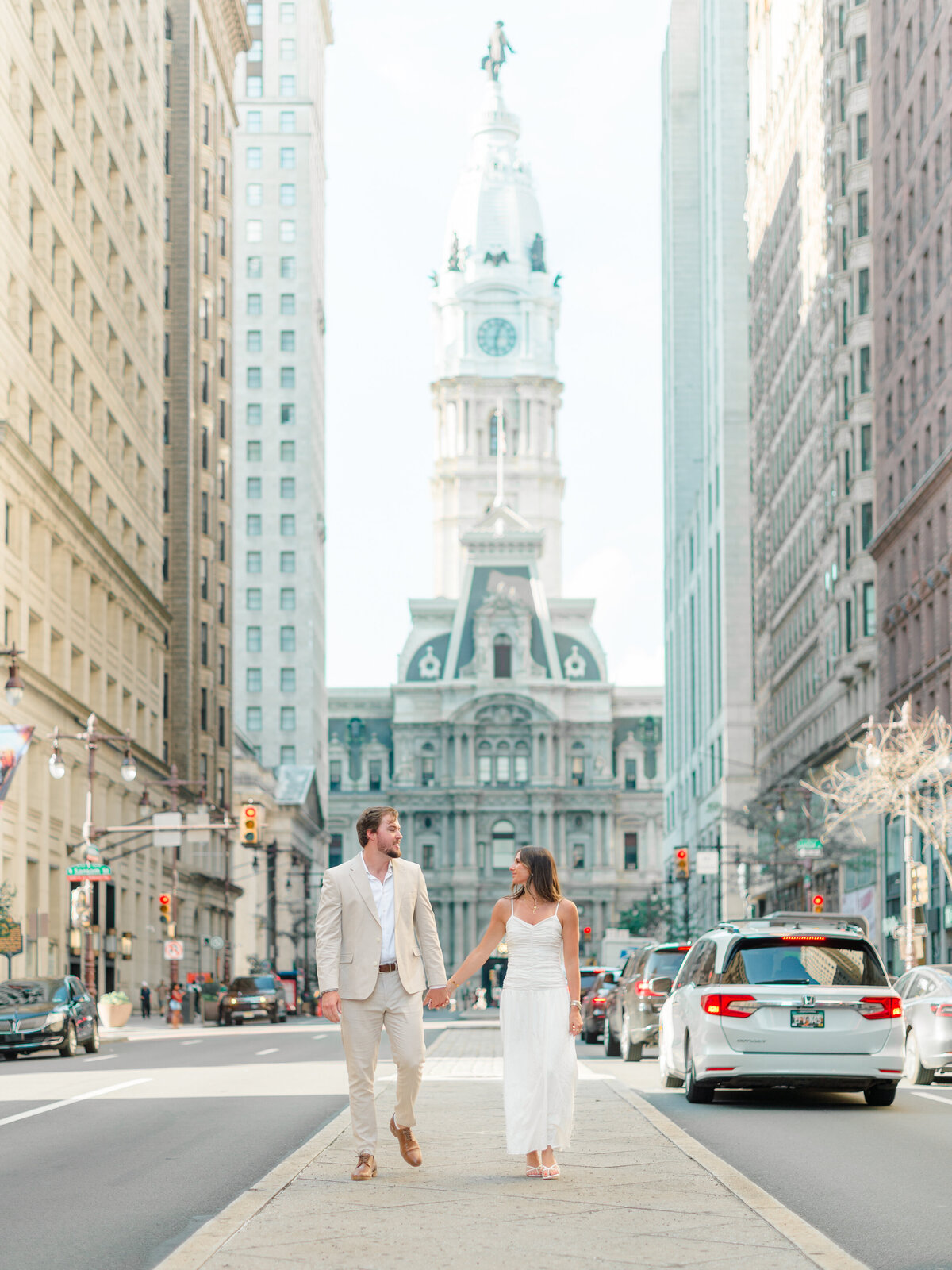 A couple walks hand in hand down the median of Broad Street in Philadelphia, with City Hall standing proudly in the distance. Dressed in light, elegant neutrals, they share a quiet moment amidst the city bustle. Captured by Elizabeth Kane Photography, a Luxury Philadelphia Wedding Photographer.