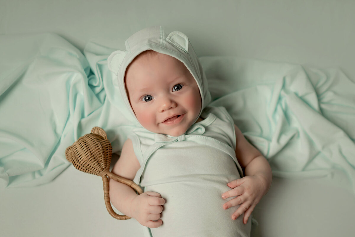 A baby in a light green outfit with a bonnet, holding a rattle lies on a light green blanket.
