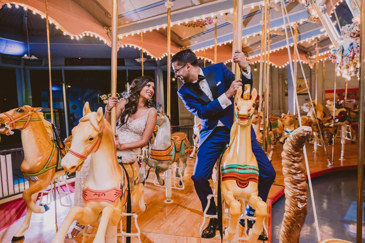 Indian bride and groom sitting on horses in a carousel carnival ride inside the please touch museum in philadelphia on their wedding day
