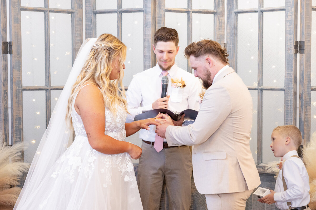 The groom gently places the wedding ring on his bride’s finger, sealing their vows in a touching exchange. The officiant stands by with a smile, while the young ring bearer watches intently. A cherished moment at Maplewood Events in Spanish Fork, Utah, beautifully photographed by Cassie Lee Photography.