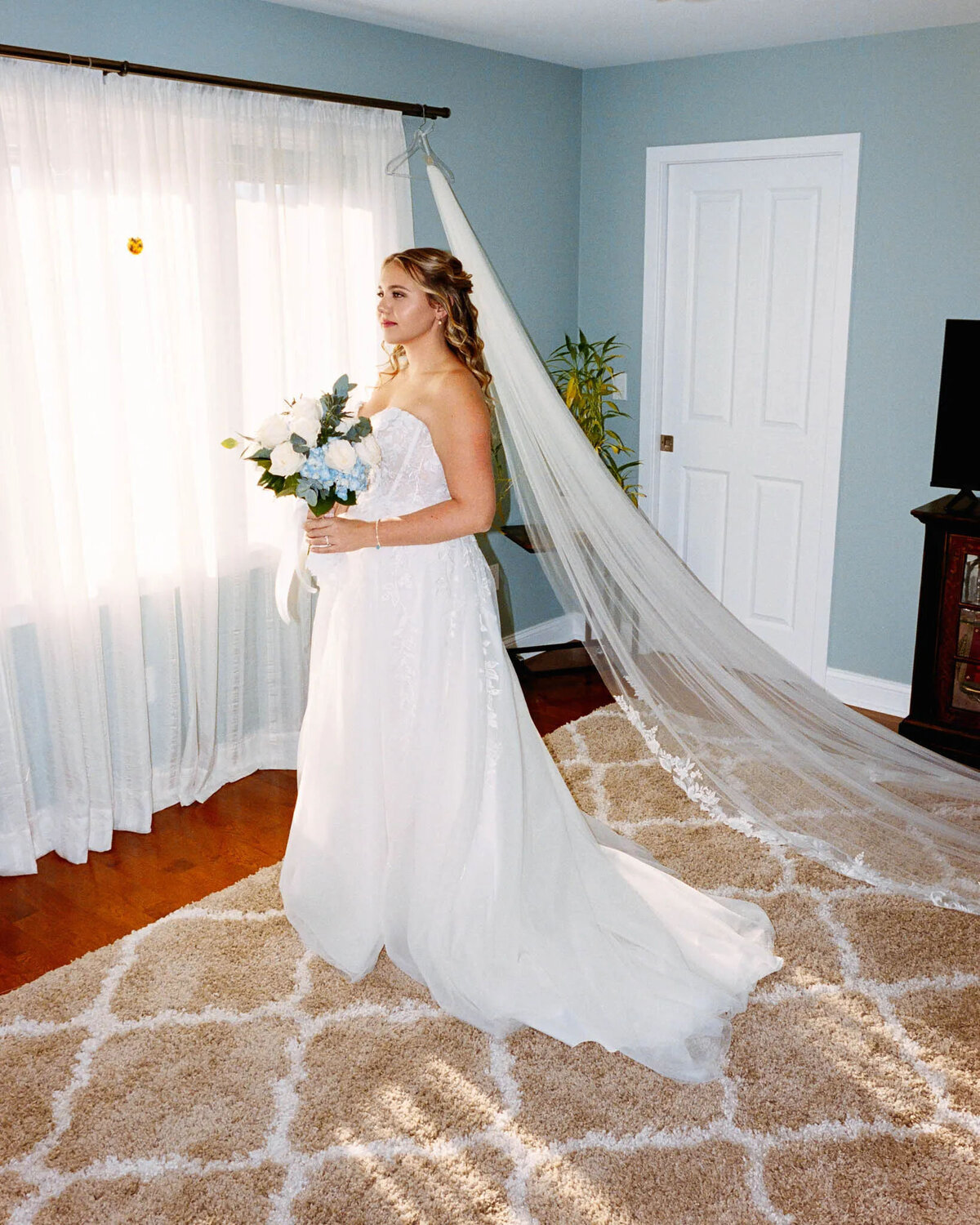 A bride in a white gown with a long veil stands on a patterned rug, holding a bouquet of flowers near a window with sheer curtains in a softly lit room, beautifully captured by an experienced NJ wedding photographer.
