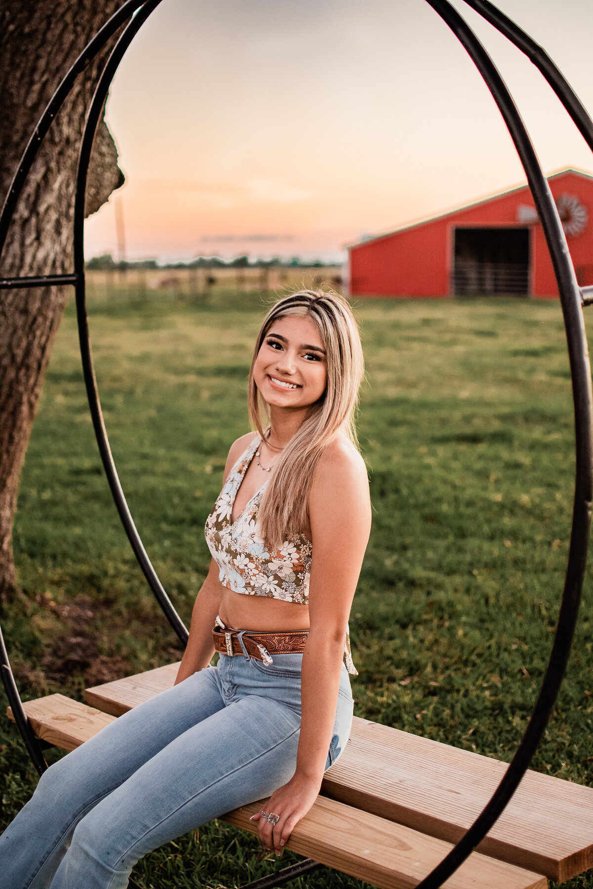 A senior wearing a floral halter top sits on a circular swing in front of a red barn.