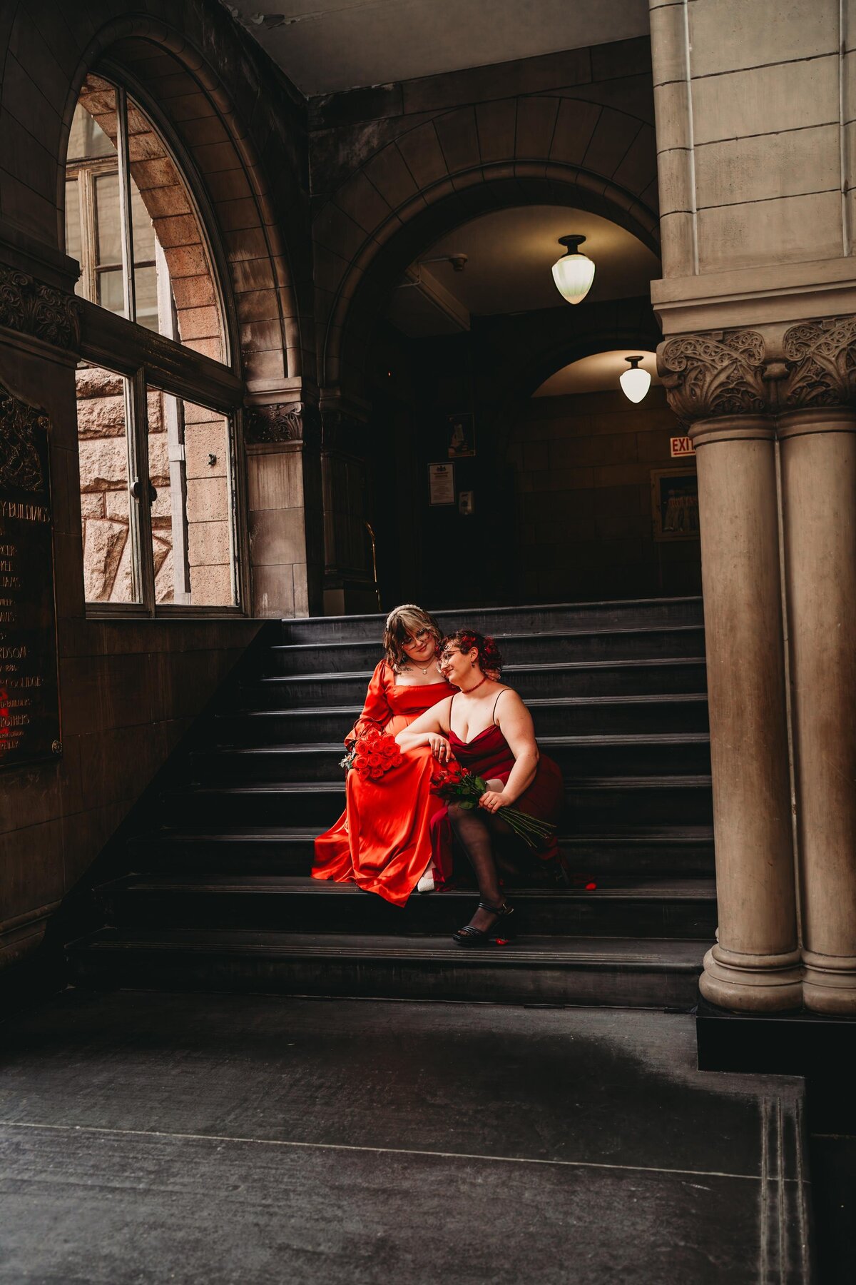 LGBTQ+ wedding portrait at Pittsburgh City Hall with a moody, cinematic feel, featuring a couple in red dresses sharing a romantic moment on historic steps.