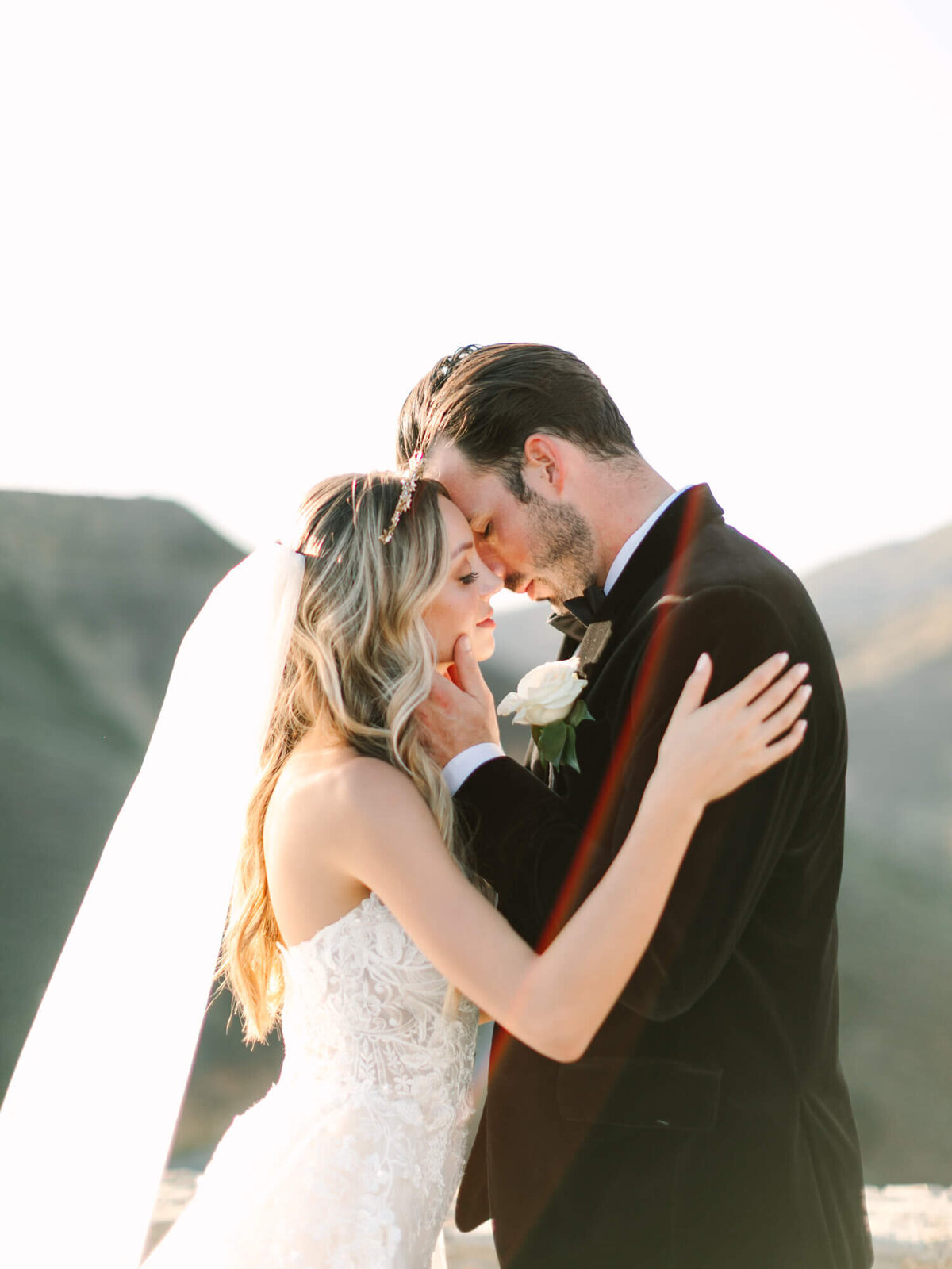 A bride and groom embrace tenderly on a sunny day, with mountains in the background.