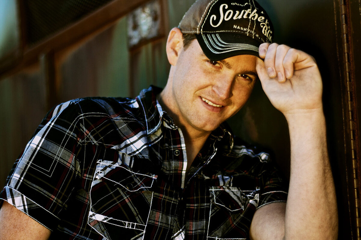 Country Music Portrait Tristan Horncastle standing by rusted old trailer holding brim of his baseball cap wearing plaid shirt