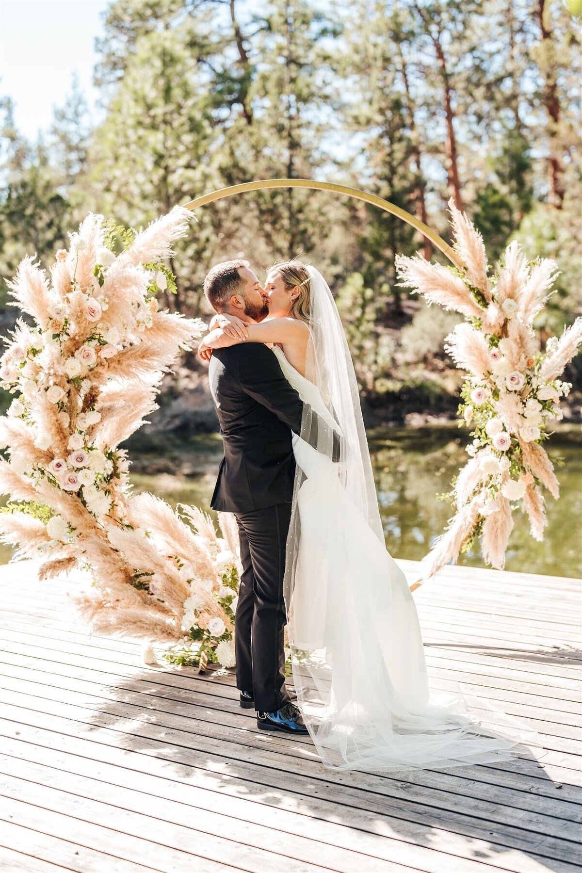 Bride and groom in front of pampas grass and roses on gold circle arch on dock at Rock Springs Ranch in Sisters Oregon