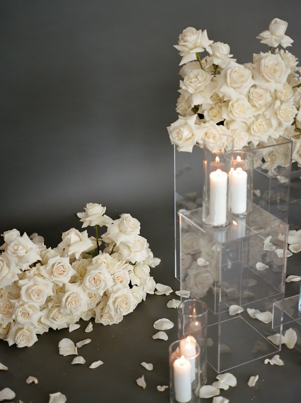 fallen petals with abundant white roses above on clear pillars, with candles and a dark gray studio background for florist brand photoshoot