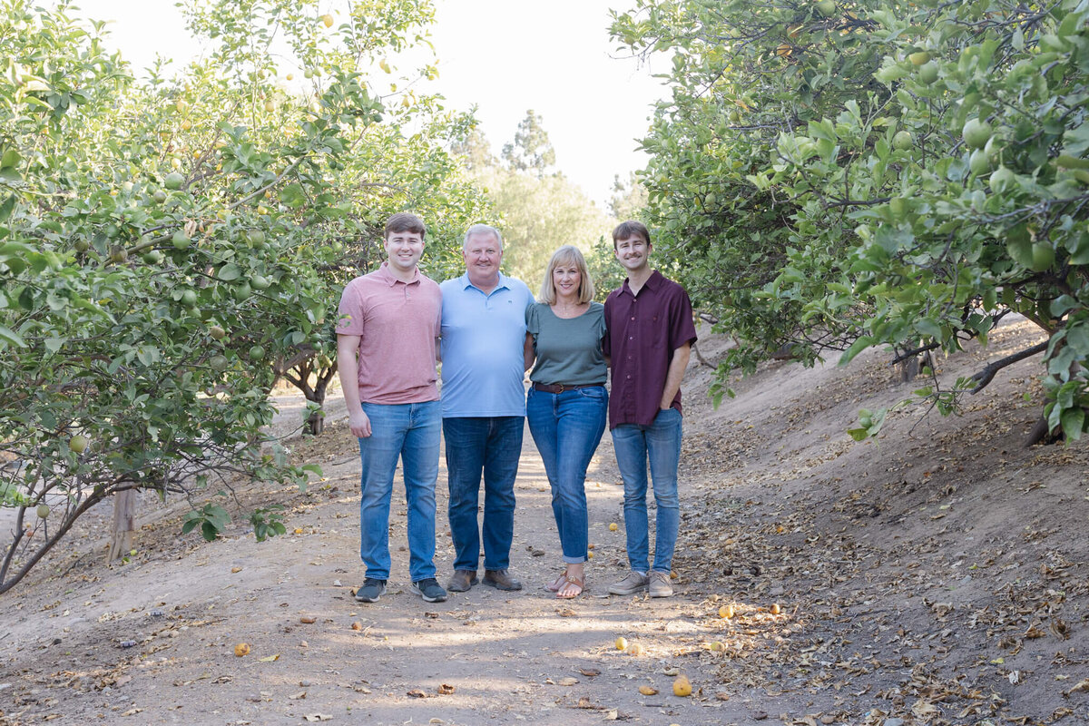 A family of four smiling at the camera in a park with green trees.