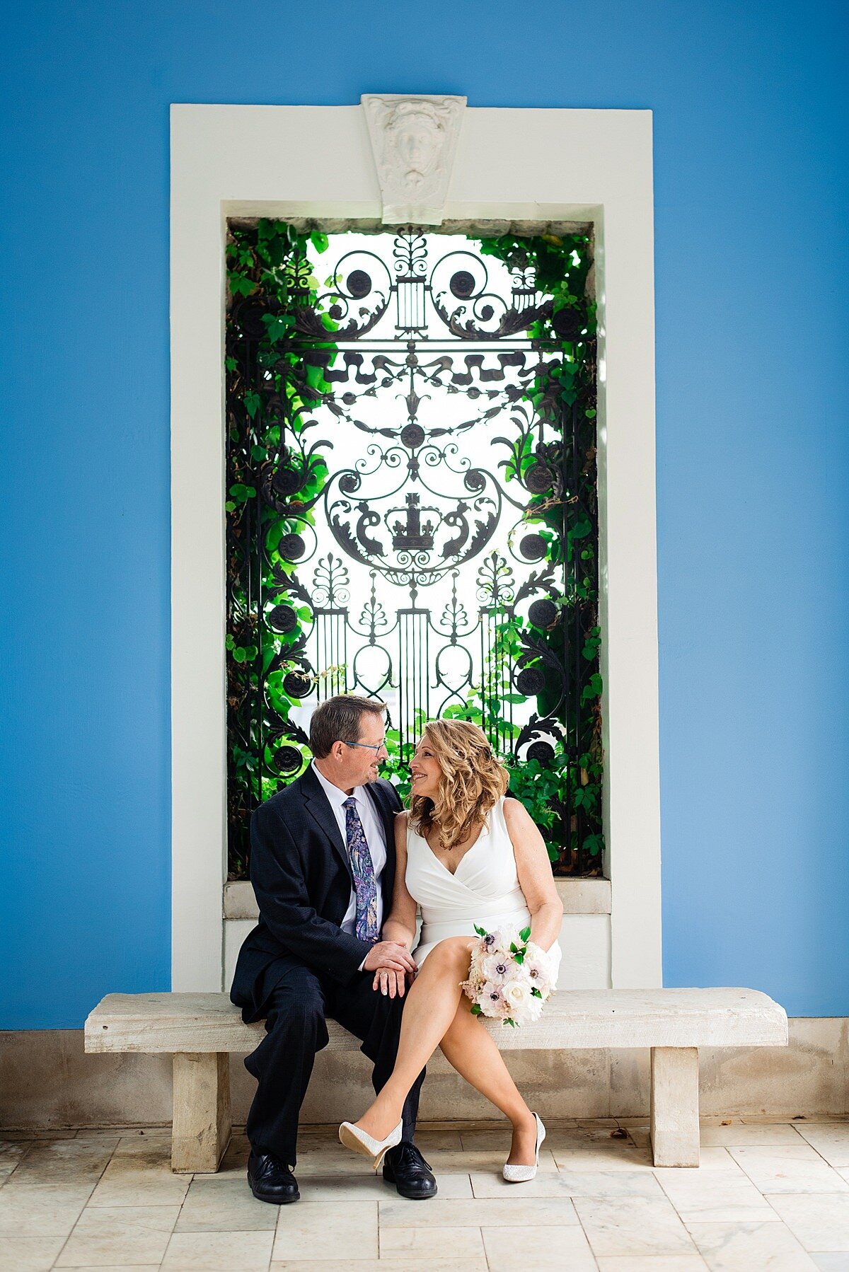 Set against a bright blue backdrop the bride and groom sit on a white stone bench in front of a window with climbing ivy and a decorative black window grate. The window is outlined in white. The bride and groom, at Cheekwood, are holding hands and looking into each other's eyes. The groom is wearing a navy suit with a white shirt and blue tie. The bride is wearing white heels with a short, fitted white dress with a v-neckline. She is holding a bouquet of white and blush flowers.