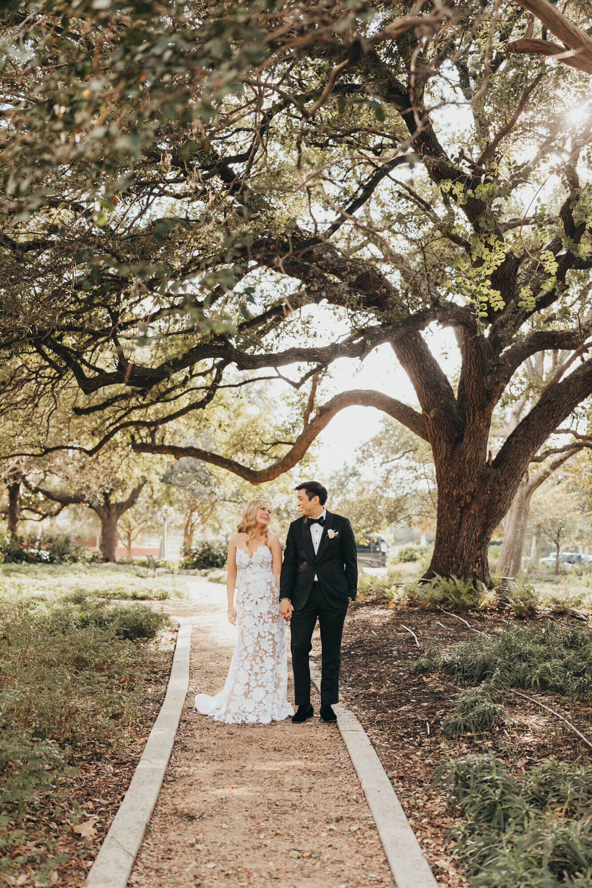 bride and groom hold hands on a trail under trees, while looking at each other.