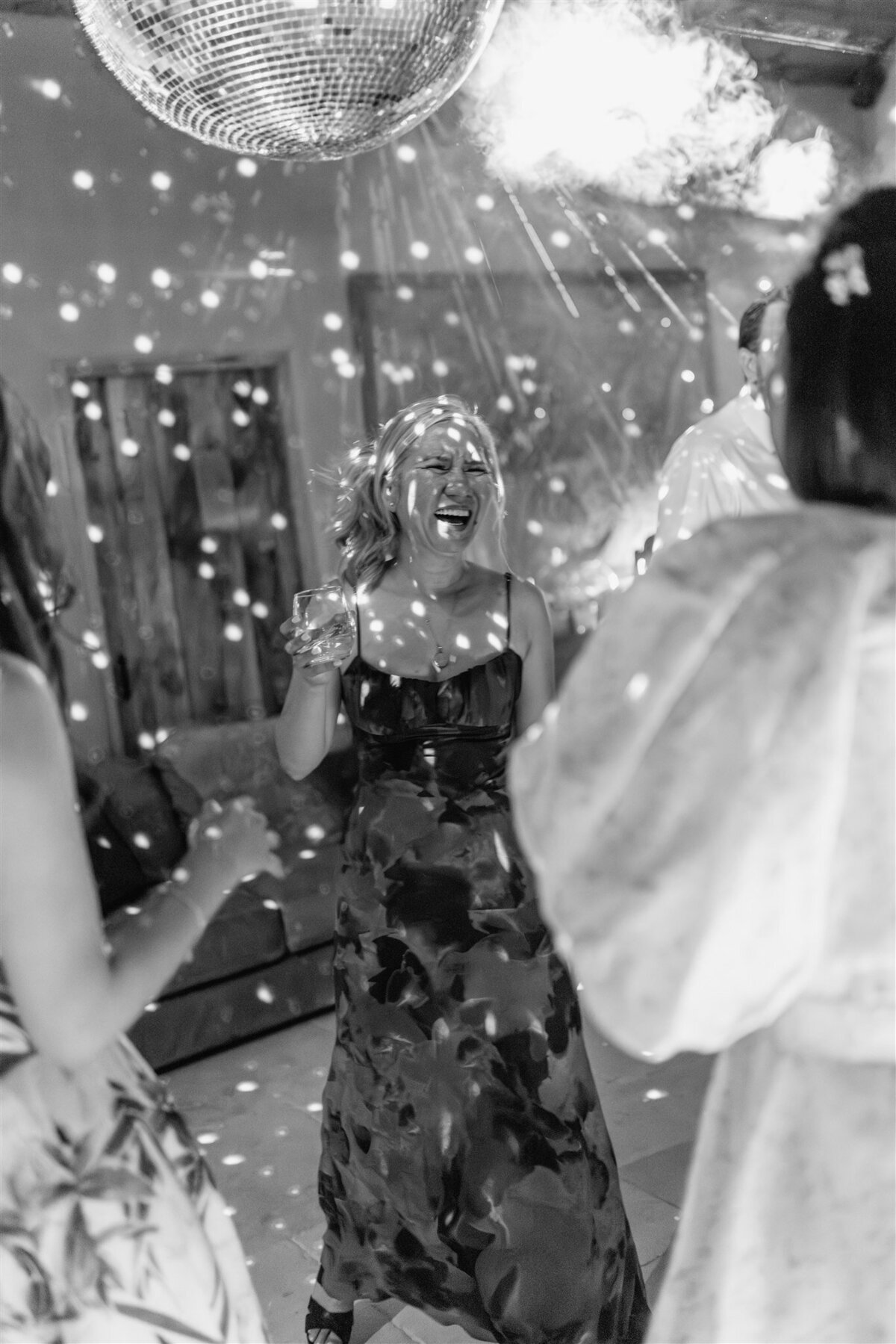 A joyful woman in a dark floral dress laughs on the dance floor under a disco ball, surrounded by friends and shimmering light reflections.