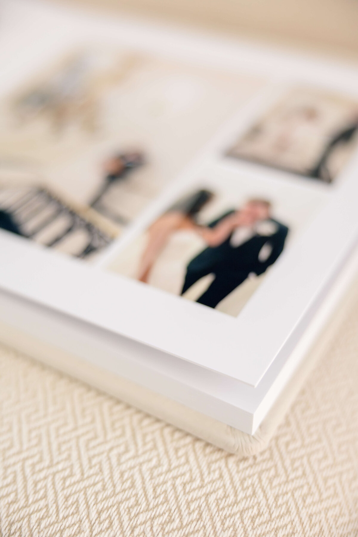 A close-up of an open photo album on a patterned surface, displaying blurred images of people in formal attire. The focus is on the edge of the album pages, emphasizing the craftsmanship and quality of the photo book.