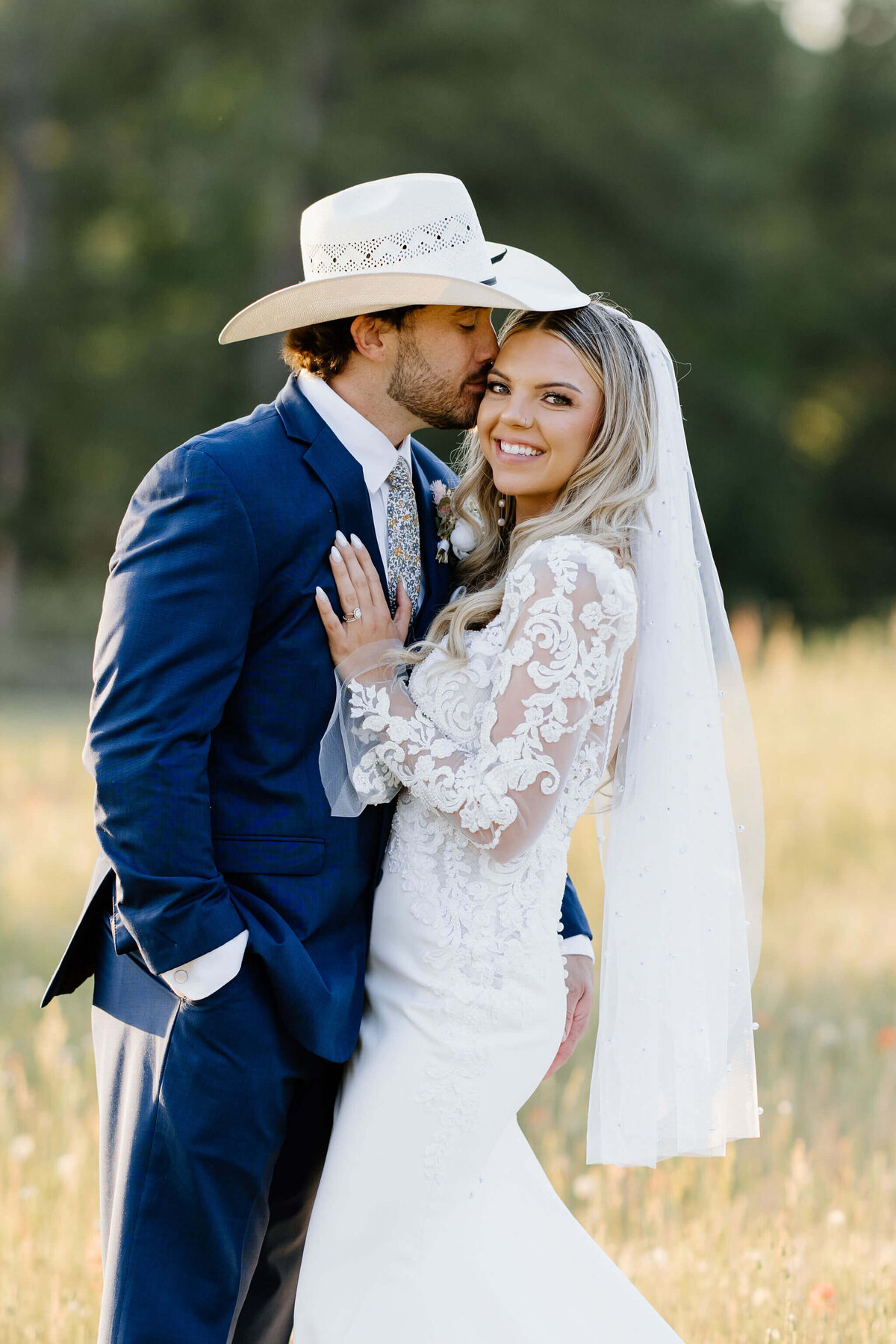 wedding day portrait of groom in navy suit and cowboy hat kissing bride in traditional lace wedding gown and pearl beaded veil