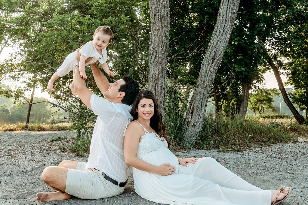 Playful outdoor maternity session with family seated by trees near water