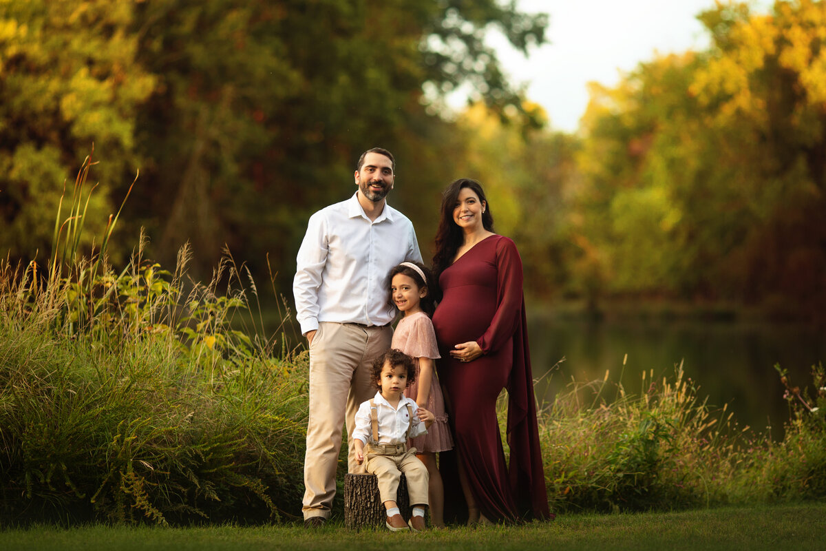 A father and his two toddler children stand in a park by a lake with pregnant mom in a maroon maternity gown