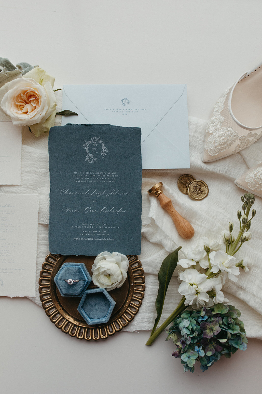 An assortment of blue and gray wedding stationery with white cursive next to a wedding ring, flowers and white wedding heels.