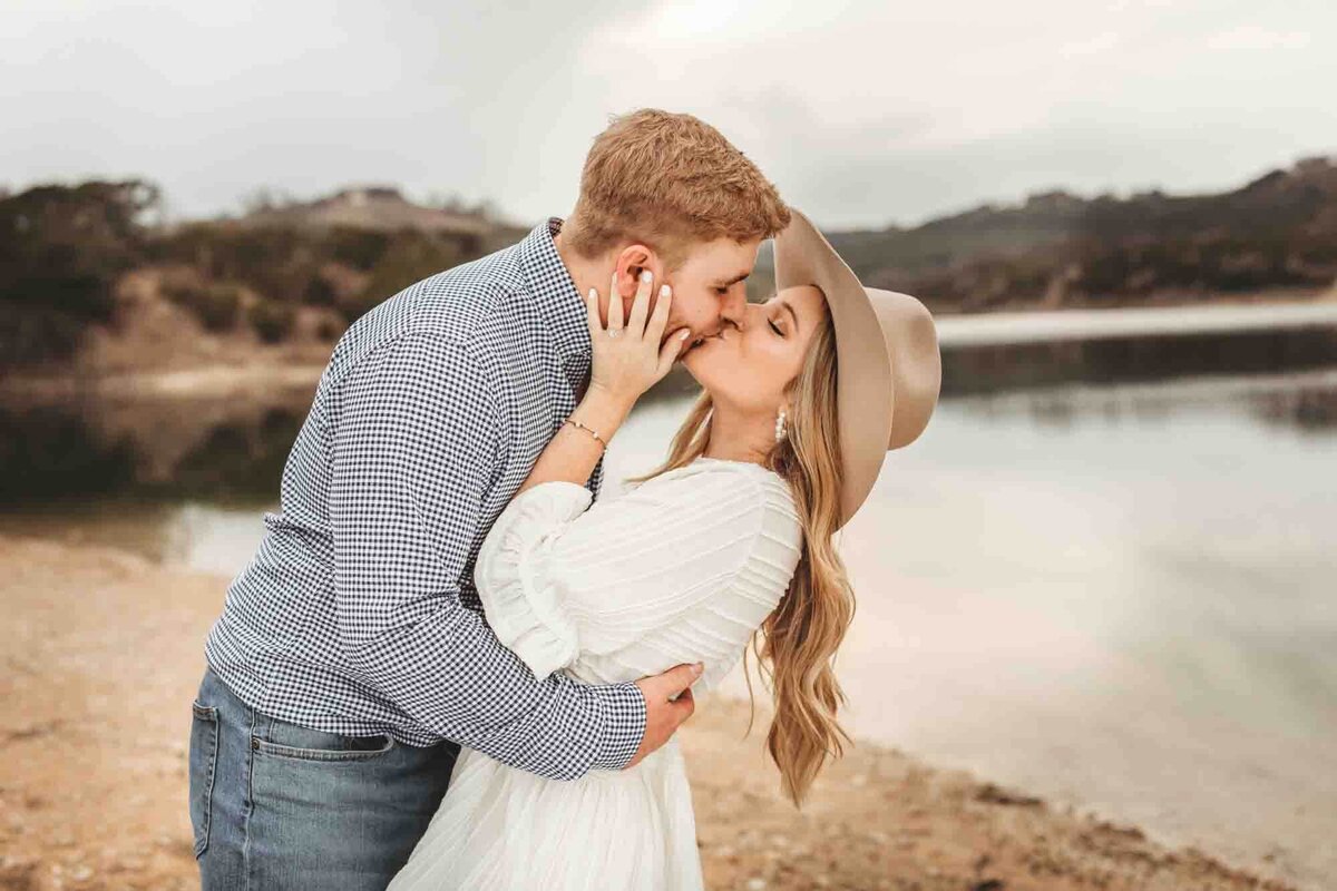 engaged couple kiss each other, leaning her over, while she wears a cowgirl hat and stands in front of a lake in Austin, Texas.