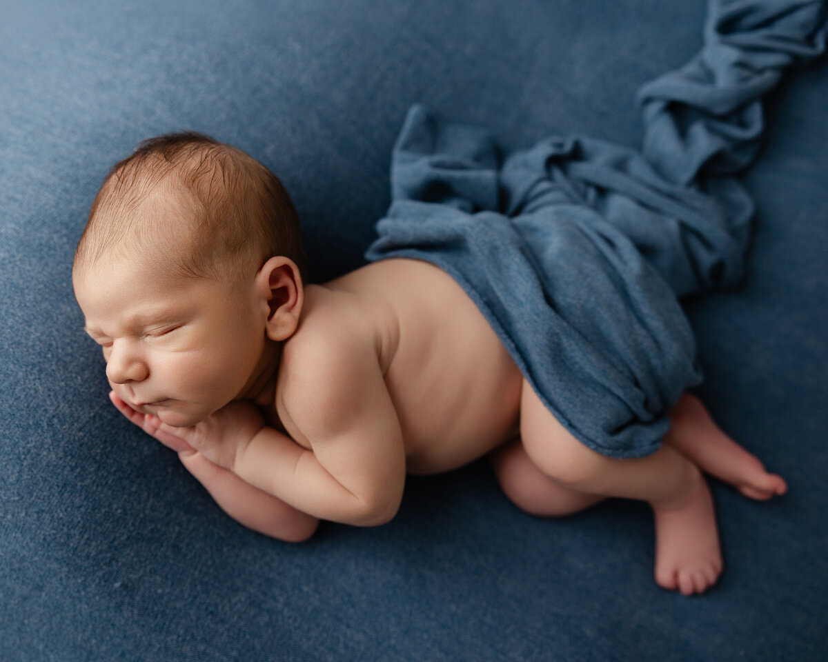 Newborn baby boy posed on a deep blue blanket with a matching wrap, sleeping with hands tucked under his chin. Captured by Inland Empire newborn photographer Kaitlyn Dawn Photography.
