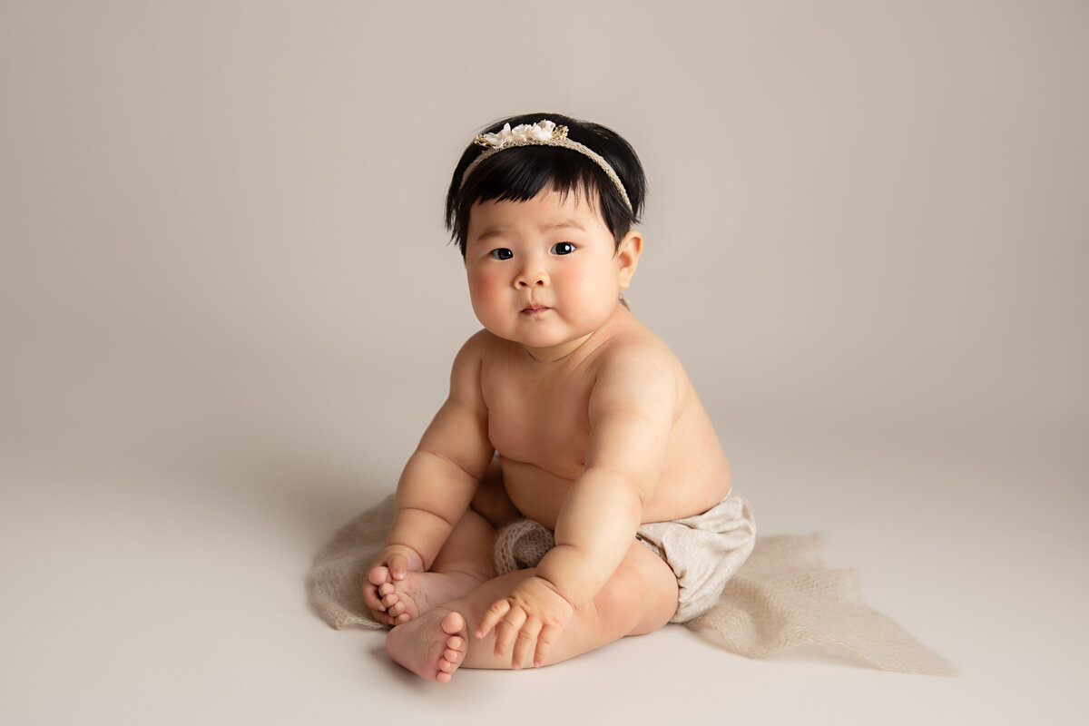 Baby sitting on a beige knitted wrap dressed in beige bloomers and a small flower headband.