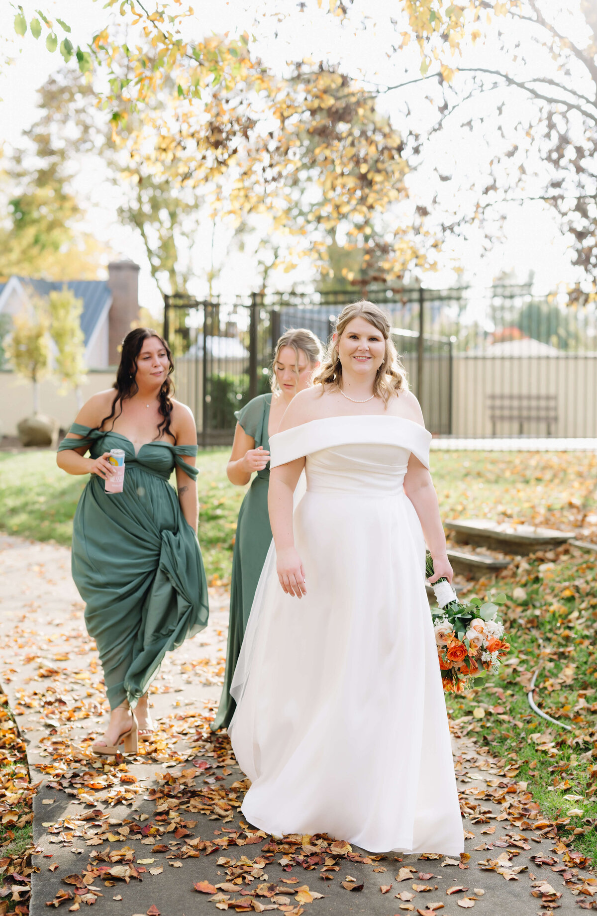 bride walking down a path outside of middleburg community center with autumn leaves at her feet as leads her bridesmaids and smiling as the sun shines through the trees