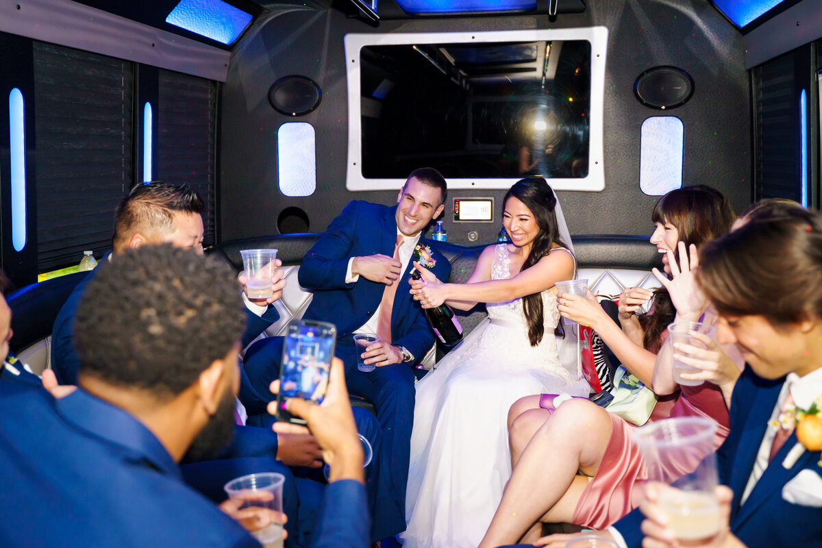 Bride laughs and pops a bottle of champagne as the wedding party looks surprised on their party bus at a wedding in Columbus, Ohio.