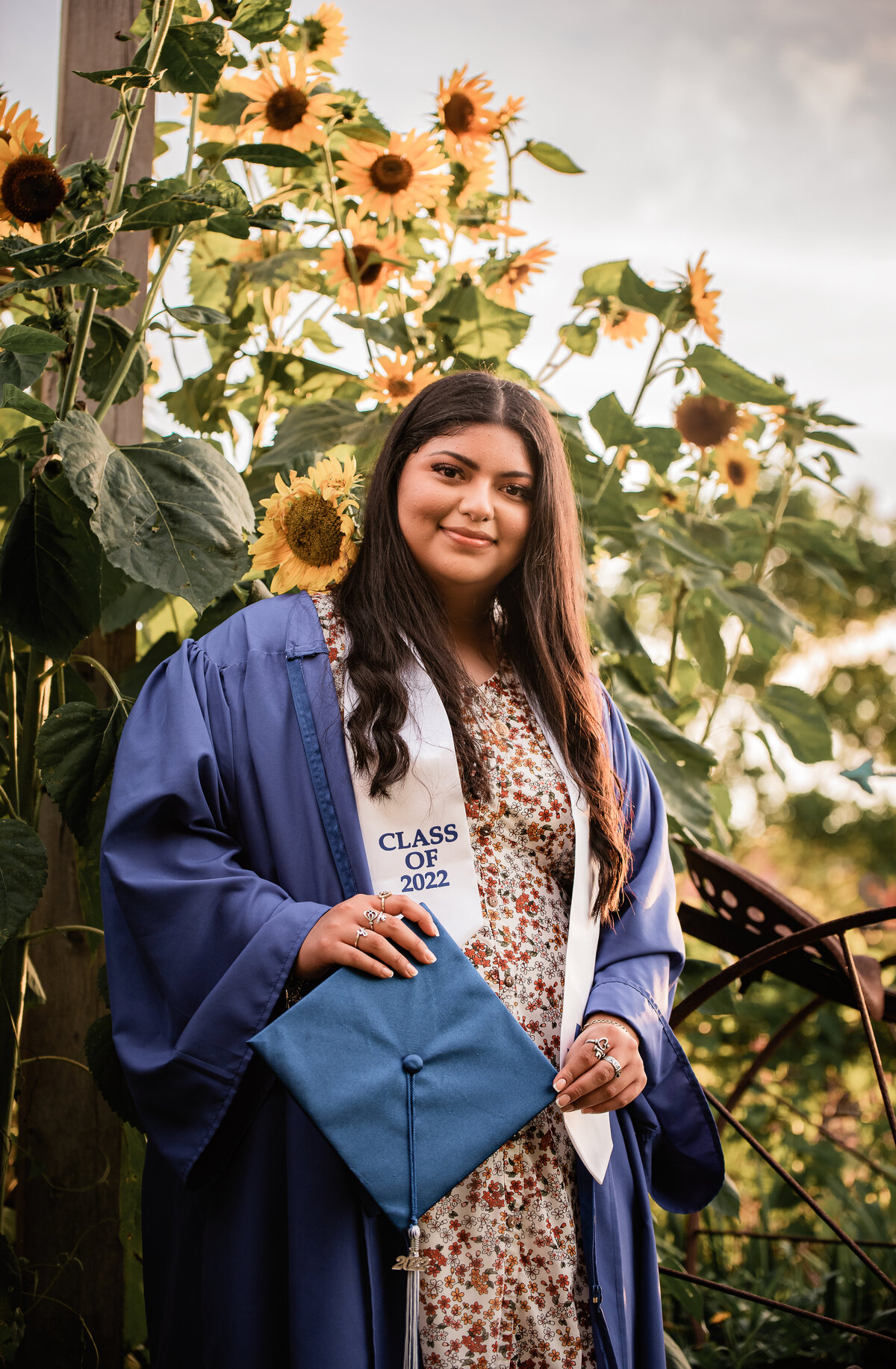 A senior wearing a blue cap and gown stands in front of sunflowers.