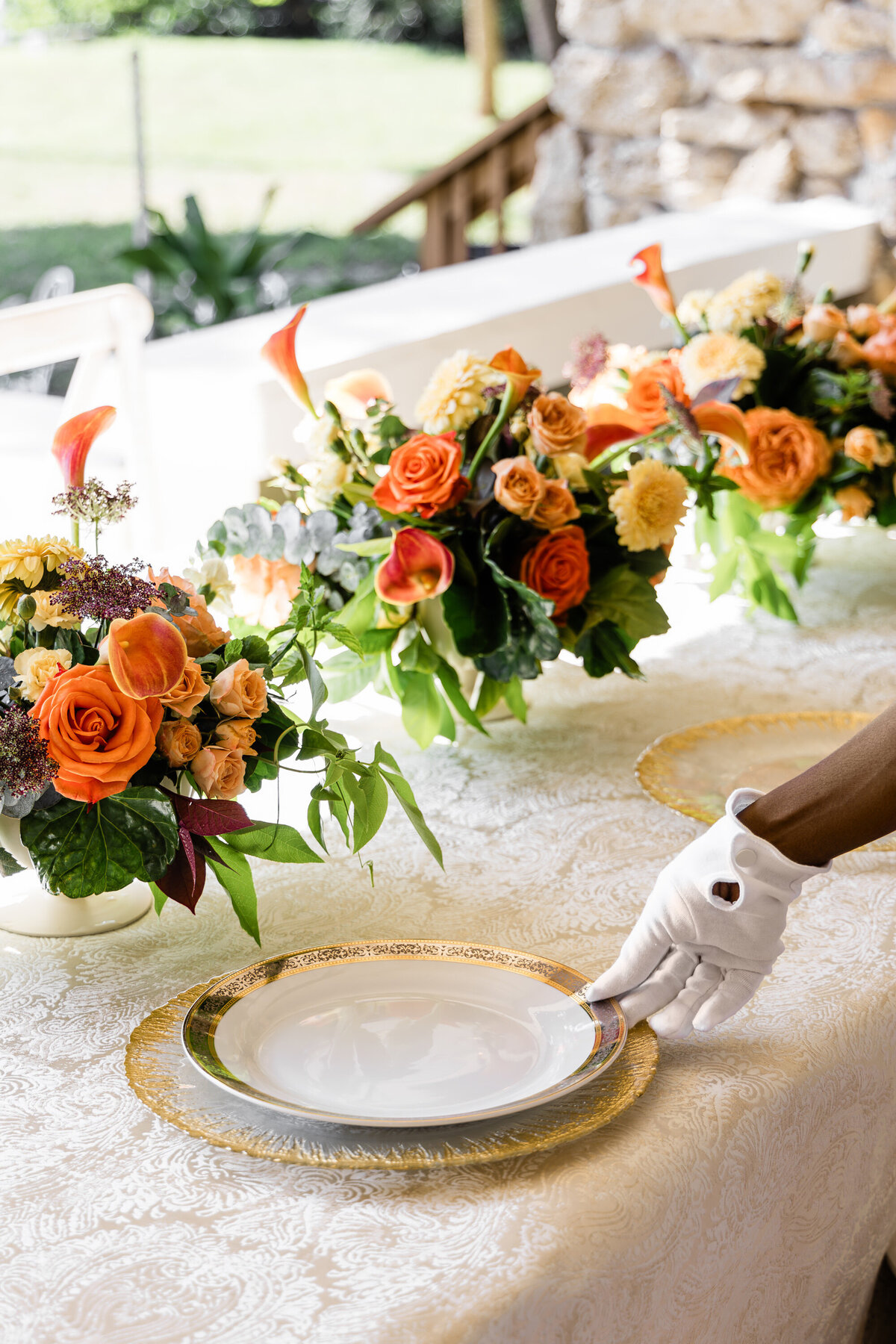 hand placing dinner plate on a glass gold sunburst charger with three orange rose and calla lily centerpieces