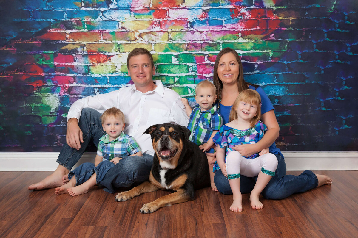 A Jacksonville family photographer captures a loving family of five posing in front of a colorful brick wall. The parents sit on the floor with two small boys and a young girl, all in matching blue outfits. Their large black and brown dog lies contentedly at their feet, completing the charming scene.