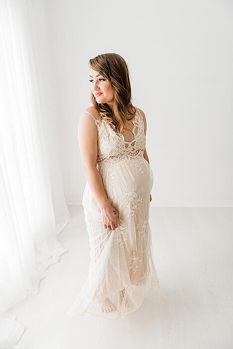 Pregnant woman in a flowy, embroidered dress stands barefoot near a sheer white curtain, softly lit from the side.