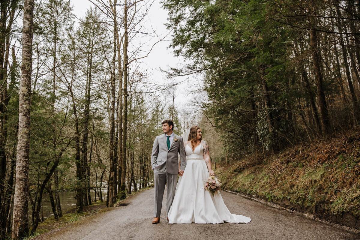 outdoor elopement pictures with bride and groom holding hands and looking opposite directions while standing on a trail in the woods in the spring