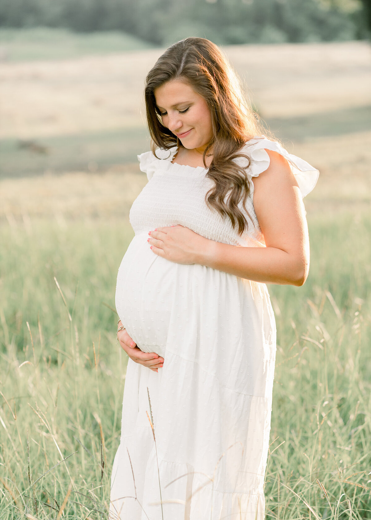 Maternity session in open green grass field. Mom to be is wearing a long white dress by central mississippi maternity photographer.