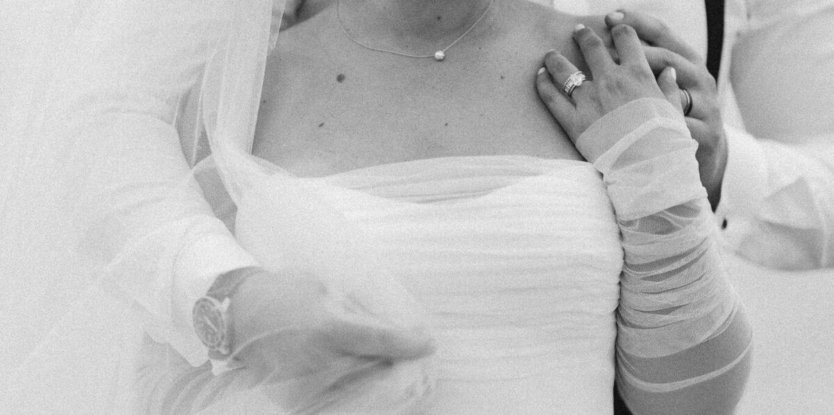 Black and white close-up of a bride's wedding details captured by Claudia Amalia Photography, showcasing her strapless gown, sheer sleeves, pearl necklace, and wedding ring resting on her partner's arm.