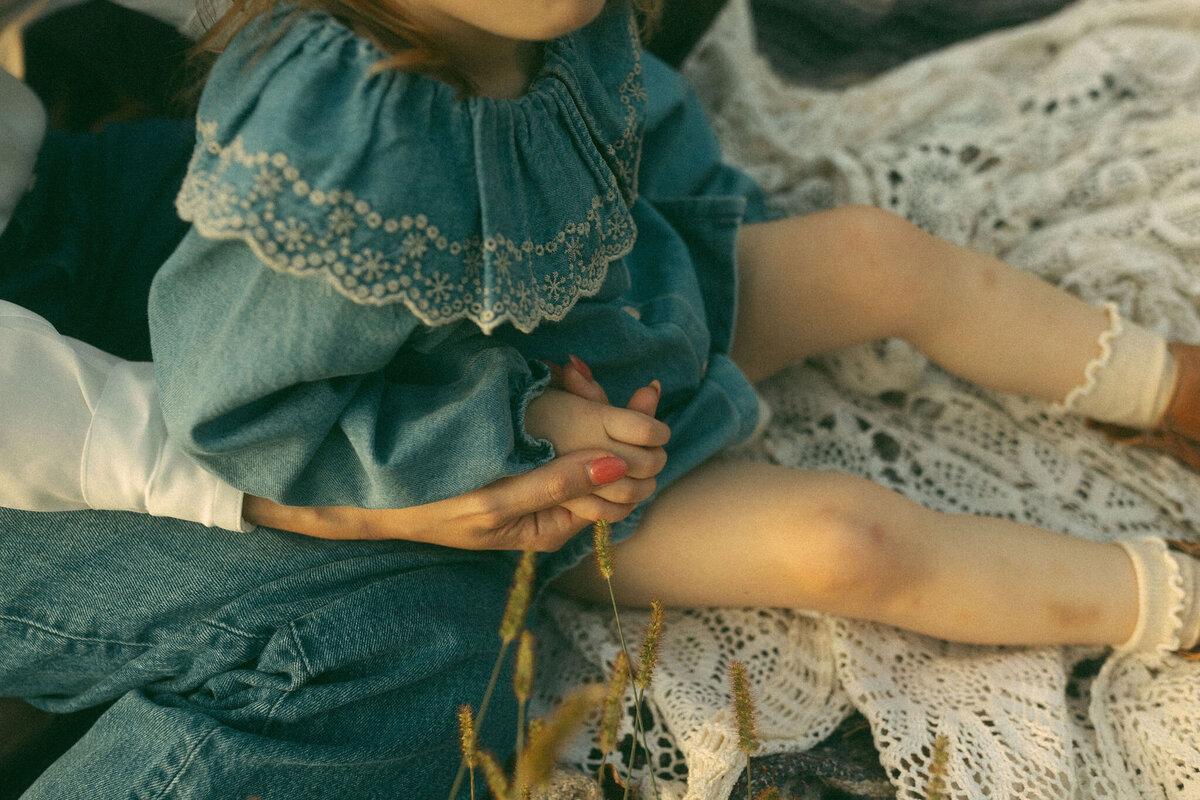 A mother sitting on a blanket with her daughter in her lap while they hold hands. 