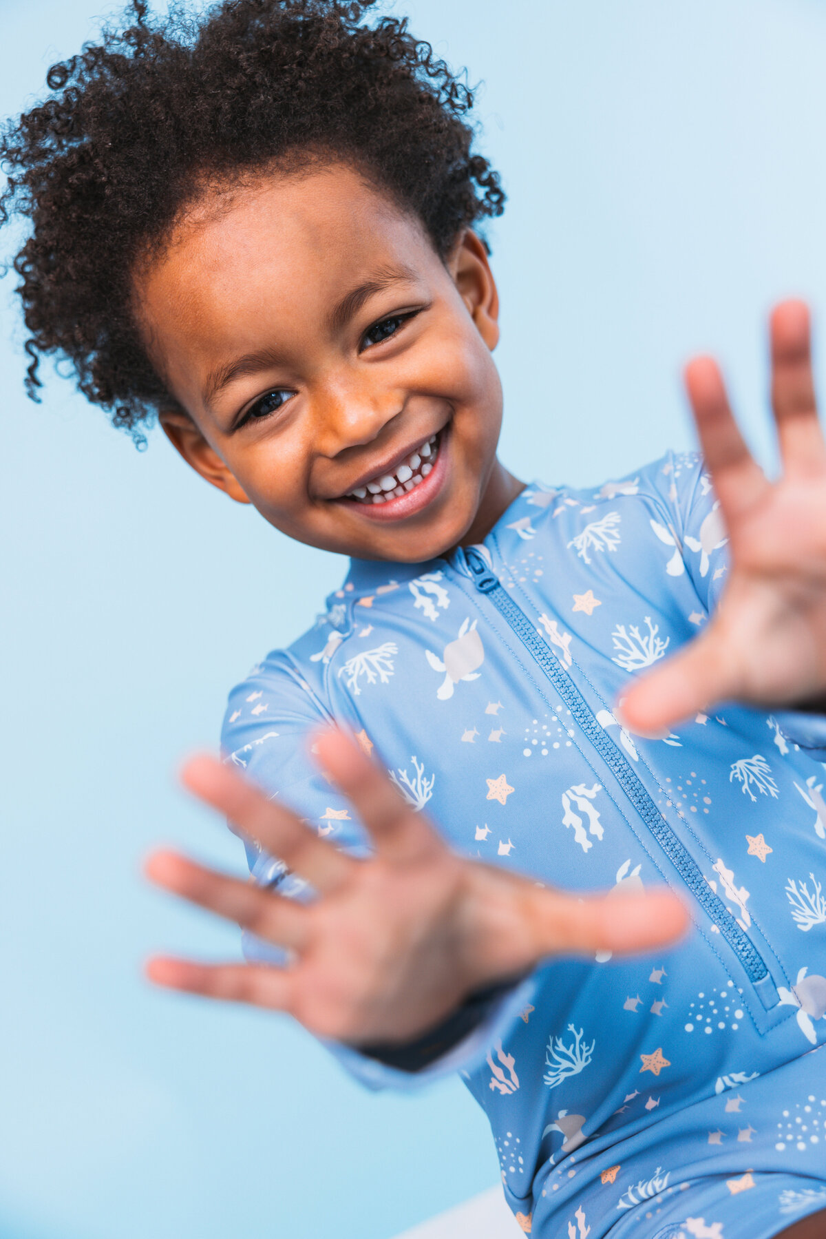 Happy child reaching towards the camera while wearing a blue sea-themed Swim Essentials swimsuit, set against a light blue background.